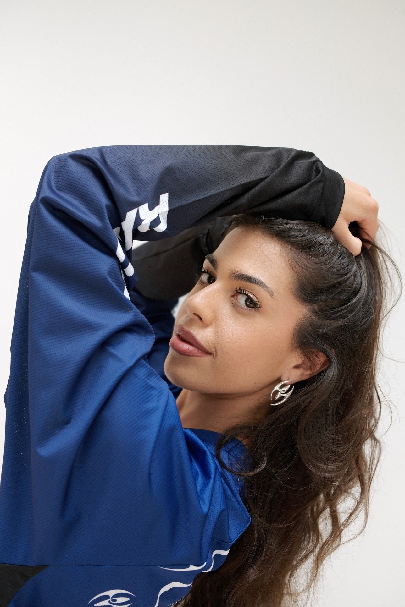 Klaudia wearing a blue and black sydocross long sleeve, posing with one arm raised against a plain light gray background showcasing a silver Sydoria earring