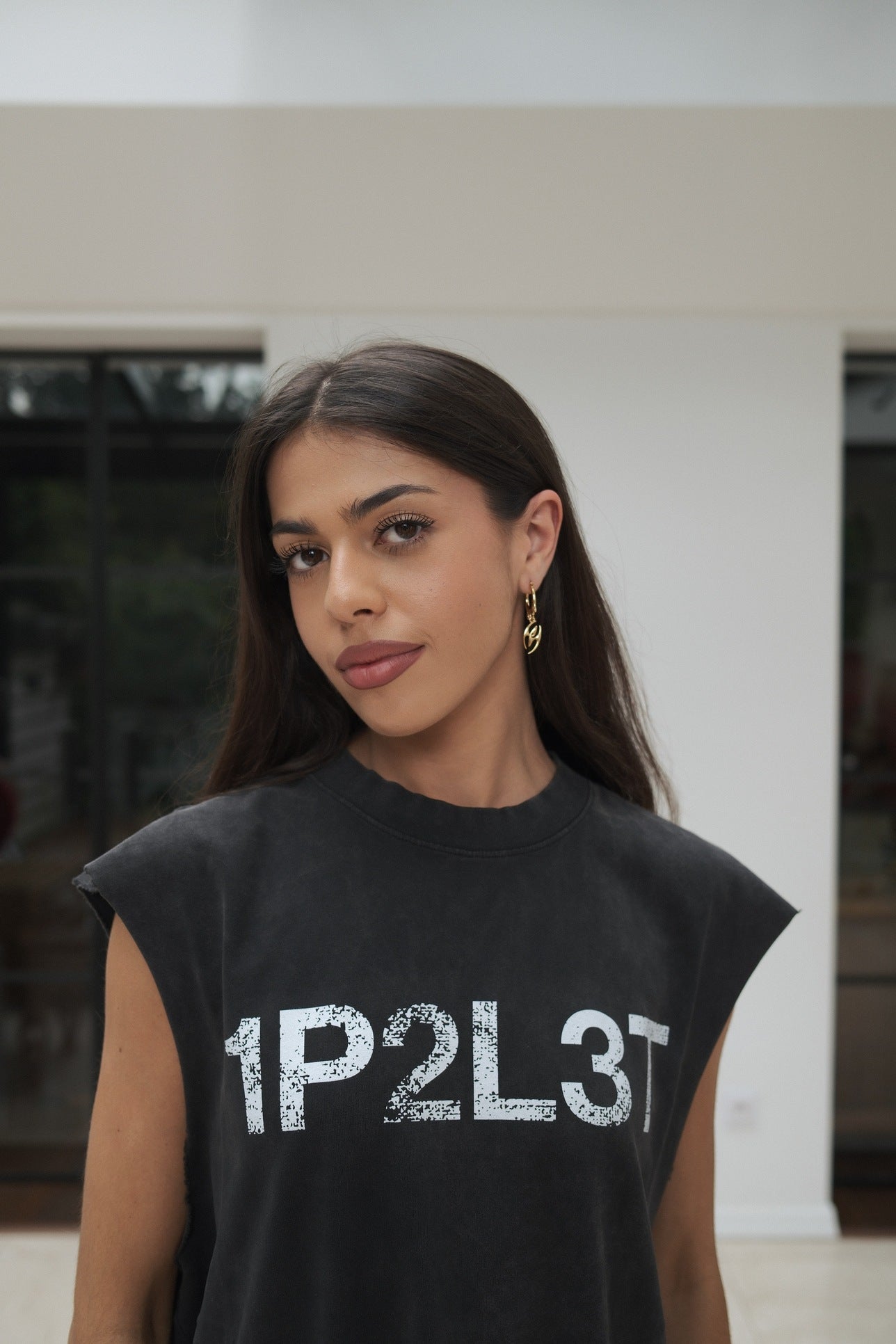 Klaudia wearing off-black, organic cotton sleeveless t-shirt with the text '1P2L3T' printed  across the chest, standing in a modern indoor setting, wearing gold Sydoria earring